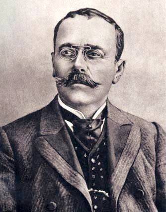 Portret alb-negru al lui Ion Luca Caragiale cu ochelari, costum și cravată