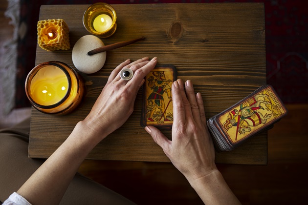 Femeie care ține în mâini o carte de Tarot, în timp ce lângă ea, pe masă, se află restul pachetului, alături de alte elemente decorative cum ar fi lumânările.