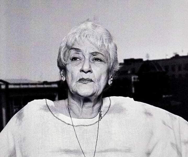 Imagine alb-negru cu Olga Tudorache cu părul alb, tunsă scurt, îmbrăcată în alb, purtând un colier lung la gât
