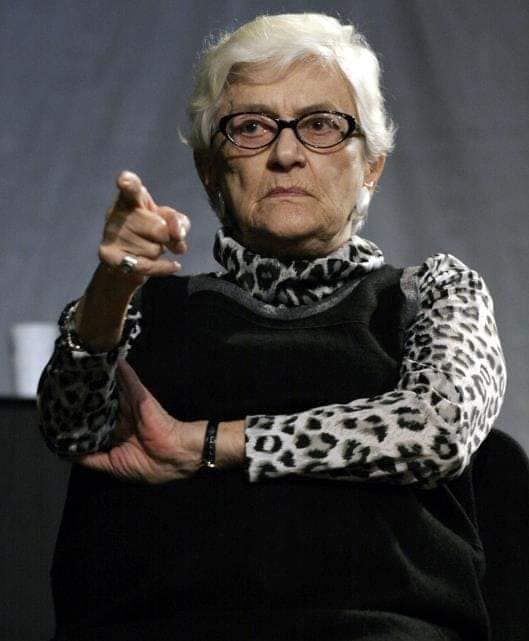 Olga Tudorache încruntată, ținând brațele încrucișate și arătând cu degetul în față, în timp ce era îmbrăcată într-o bluză cu imprimeu animal print, peste care avea un sarafan negru.