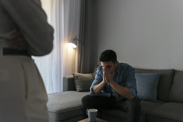 Femeie care stă în picioare în fața partenerului așezat pe canapea, în timp ce așteaptă ca el să spună ceva.