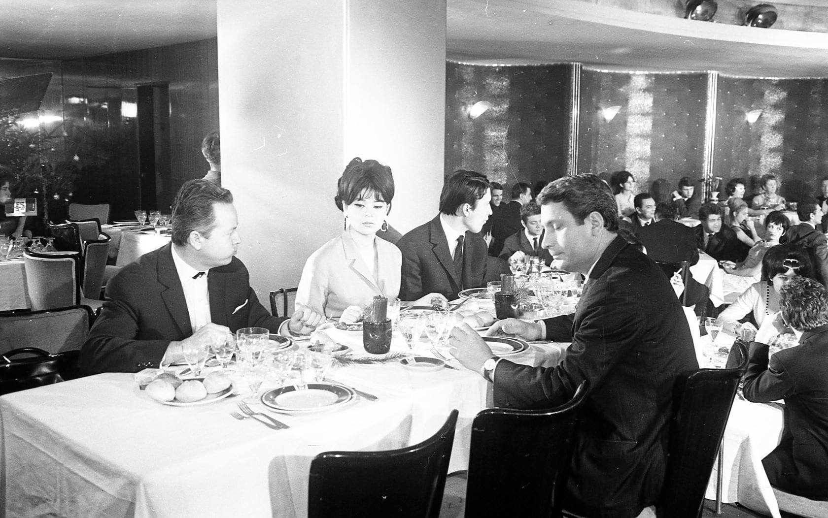 Imagine alb-negru cu Cristina Stamate în tinereșe, așezată la o masă, înconjurată de bărbați