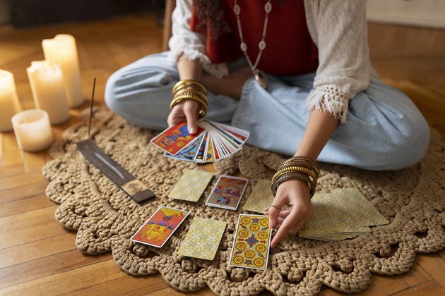 Femeie care stă pe podea și ghicește viitorul în cărțile de tarot, așezate pe covor, lângă alte elemente de magie neagră