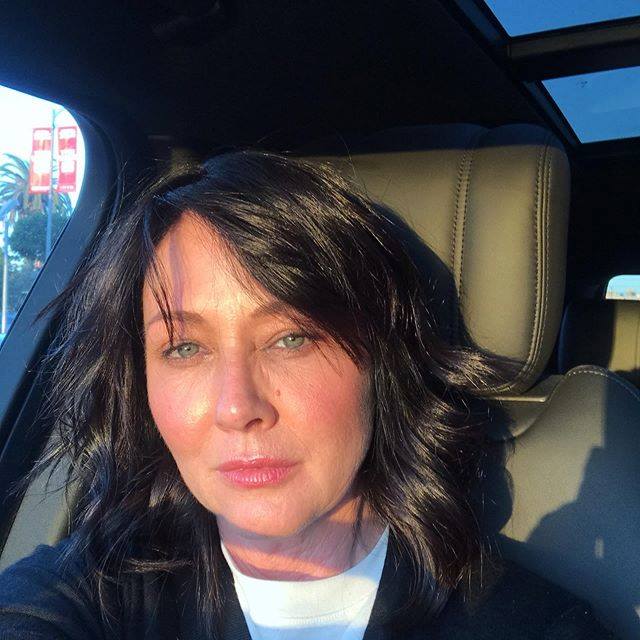 Shannen Doherty pe scaunul mașinii, în lumina soarelui, îmbrăcată în alb și negru