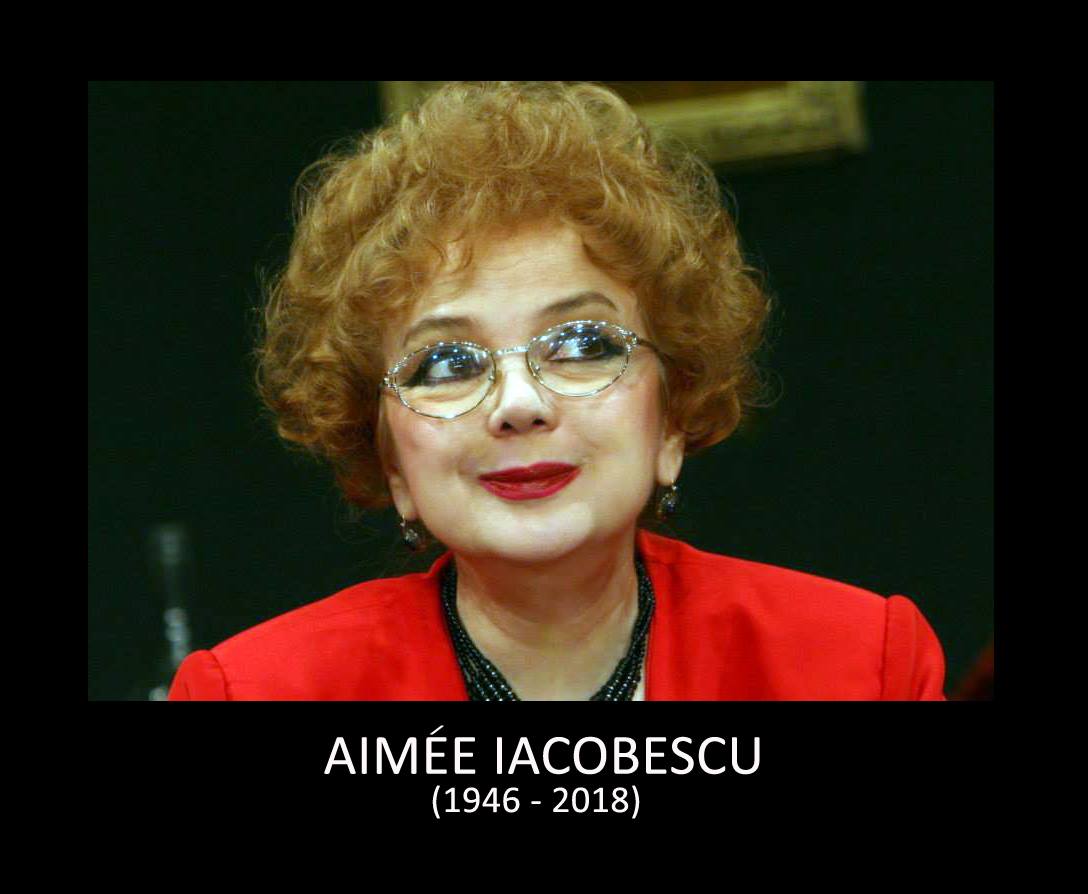 Aimée Iacobescu într-un sacou roșu, cu ochelari de vedere și mărgele închise la culoare