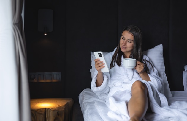 Femeie în halat alb pufos, care stă în pat, în camera de hotel și savurează o cafea, în timp ce se uită în telefon