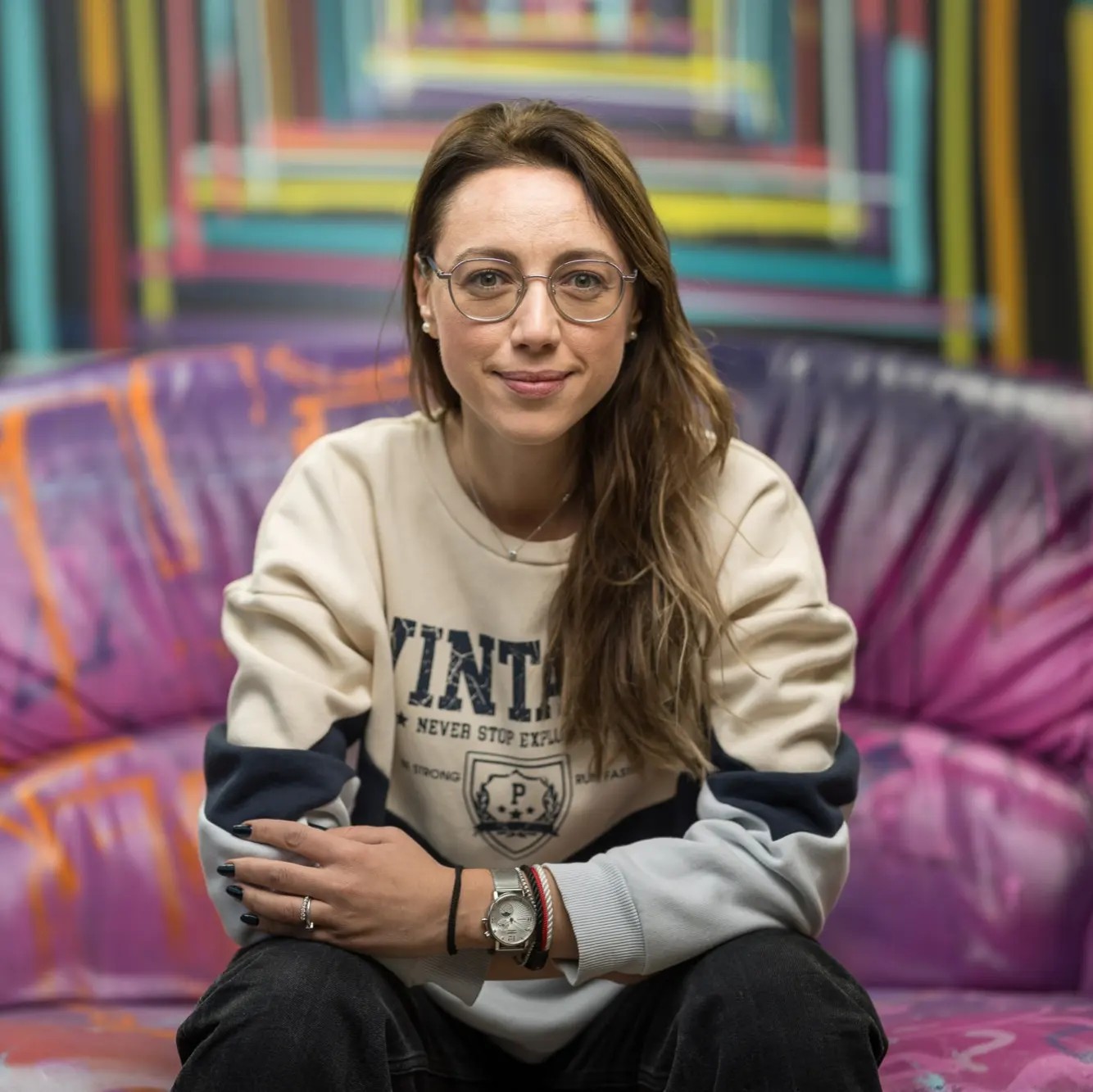 Andreea Preda stând pe o canapea mov cu portocaliu, în timp ce este îmbrăcată într-un hanorac bej cu albastru închis și poartă ochelari de vedere