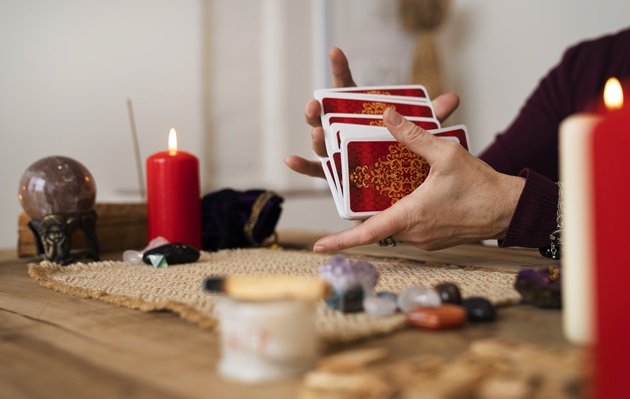 Mâinile unei femei care amestecă toate cărțile din pachetul de Tarot, în timp ce are o lumânare roșie, aprinsă, pe masă