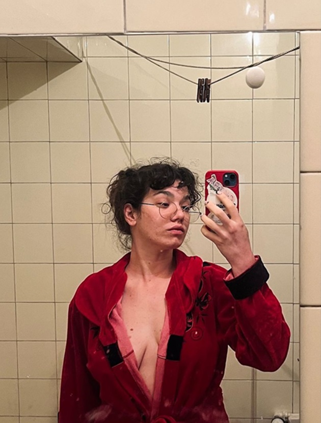 Cabiria Morgenstern în pijamale, cu părul strâns, în timp ce își face poză în oglindă și poartă ochelari