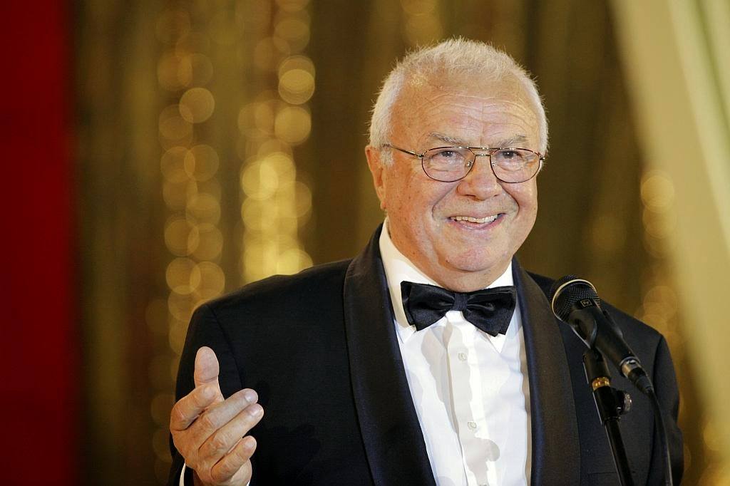 Alexandru Arșinel pe scenă, cu microfonul în față, în costum negru și cămașă albă, accesorizată cu papion negru