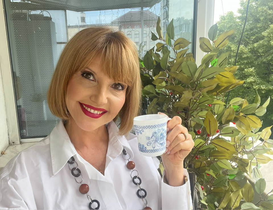 Marina Almășan, zâmbitoare pe balcon, stând lângă o plantă și ținând în mână o ceașcă de ceai