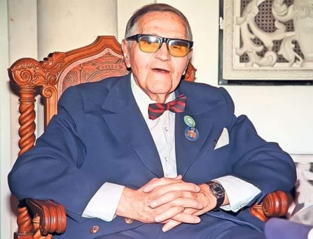 Gică Petrescu în ultima parte a vieții, stând pe un scaun de lemn, îmbrăcat în costum albastru închis, cu ochelarii la ochi și cu mâinile încrucișate