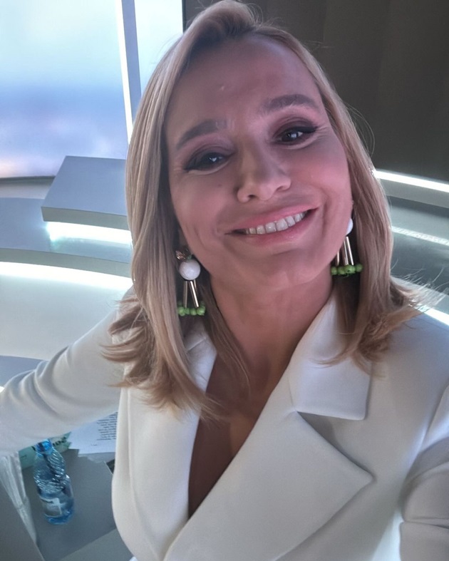 Andreea Esca făcându-și un selfie la pupitrul știrilor, în timp ce poartă un sacou alb și cercei voluminoși în formă de triunghi
