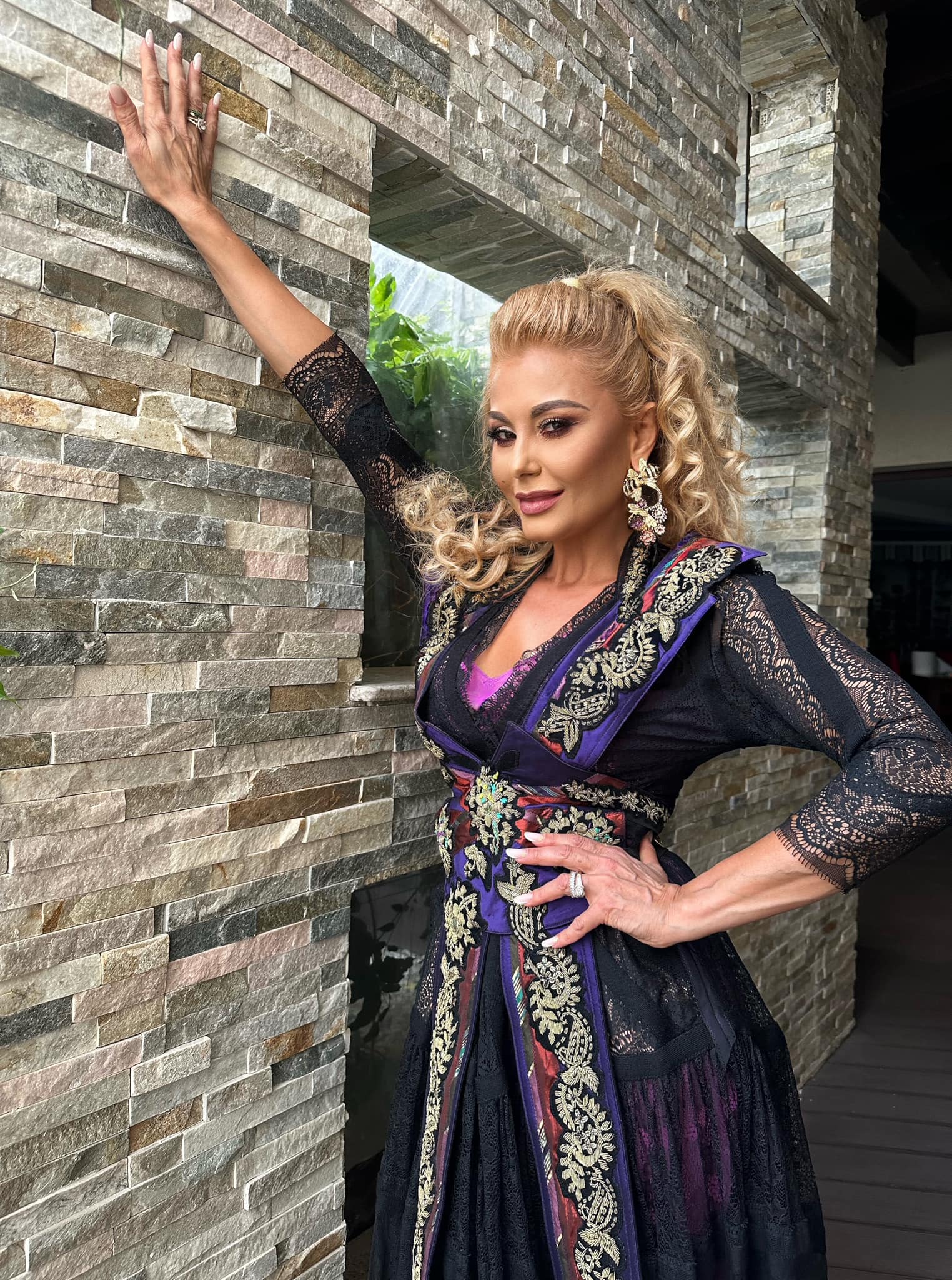 Emilia Ghinescu îmbrăcată într-o rochie neagră din dantelă, cu părul ondulat, sprijinindu-se de un perete