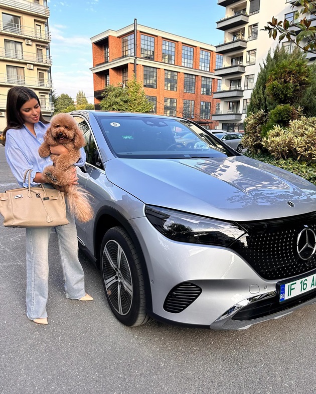 Ileana Lazariuc ținând un cățel în brațe și o geantă pe mână, în timp ce stă lângă o mașină electrică de lux