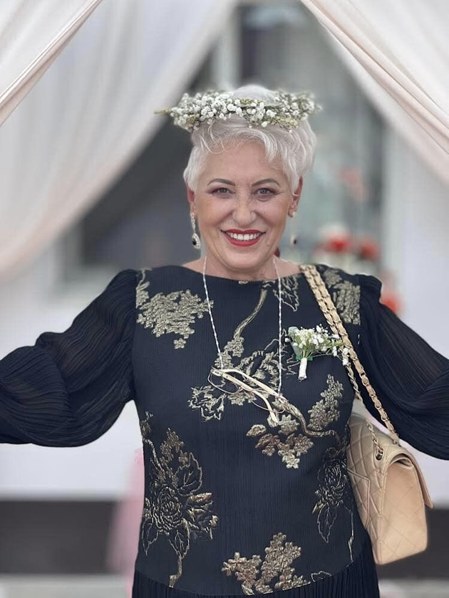 Lidia Fecioru la o nuntă, cu floare în piept și coroniță pe cap, îmbrăcată într-o rochie neagră cu imprimeu floral auriu