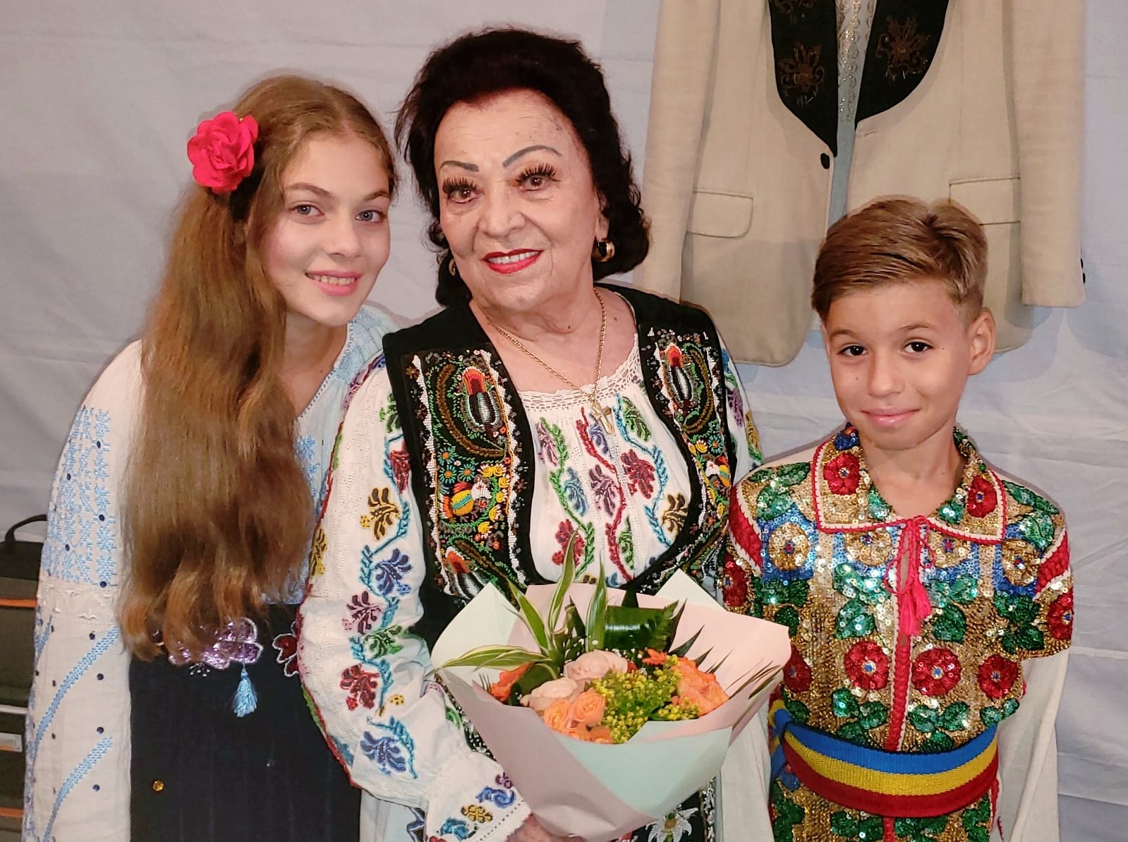 Maria Ciobanu în costum popular, cu un buchet de flori în mână, alături de doi dintre nepoții ei, care îi calcă pe urme