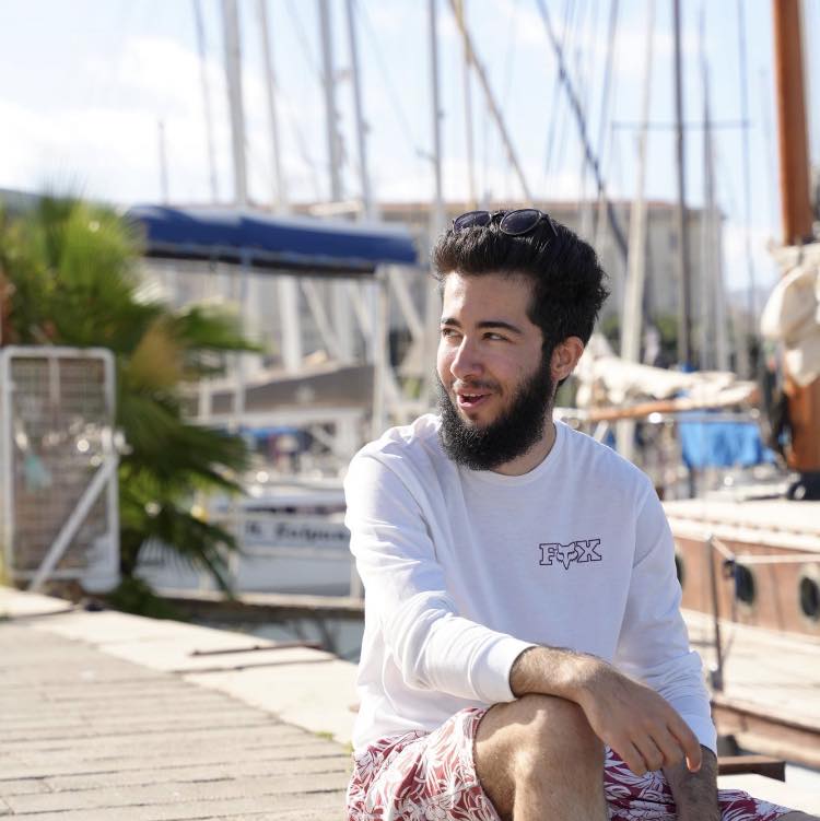Arian Mirzarafie-Ahi stând într-un port, privind într-o parte, îmbrăcat în bluză albă și pantaloni colorați