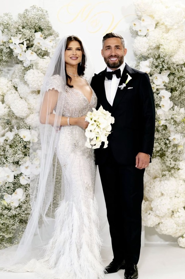 Monica Bîrlădeanu și Valeriu Gheorghiță îmbrăcați în mire și mireasă, lângă panoul foto din flori, la nunta lor