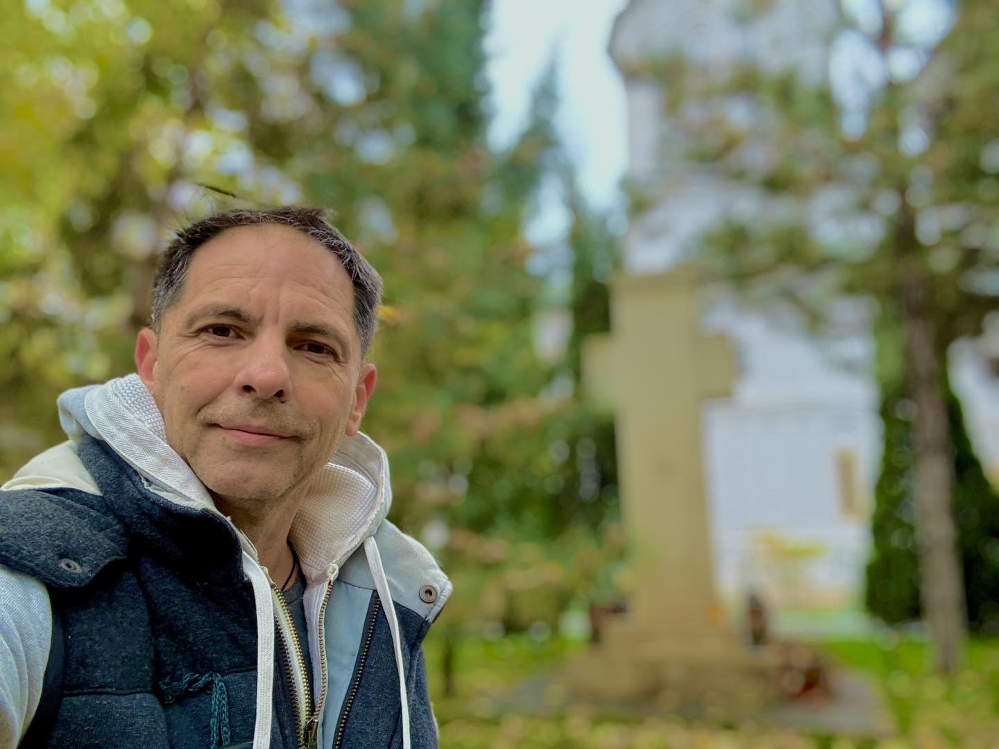 Dan Negru îmbrăcat într-o vestă de culoare albastru închis, făcându-și un selfie lângă un monument în formă de cruce