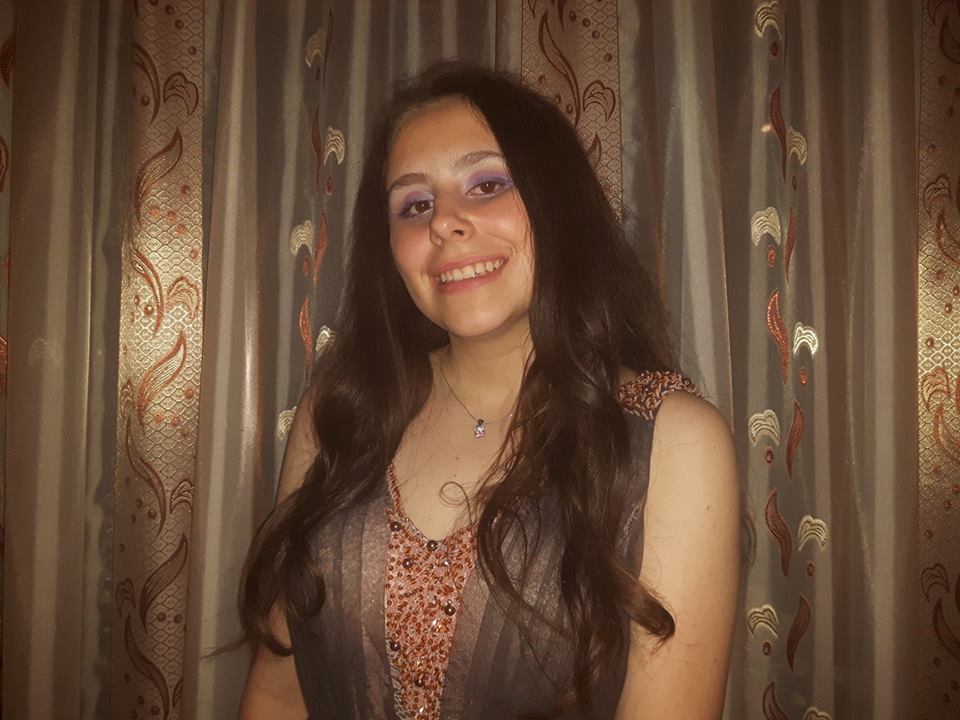Cătălina Toma într-o rochie roz elegantă, stând lângă o draperie maro cu auriu