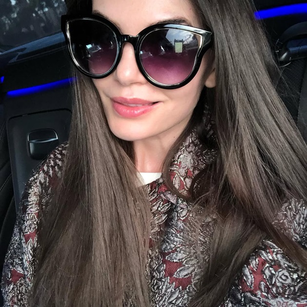 Monica Gabor făcându-și un selfie în mașină, purtând ochelari de soare și un sacou maro cu flori argintii