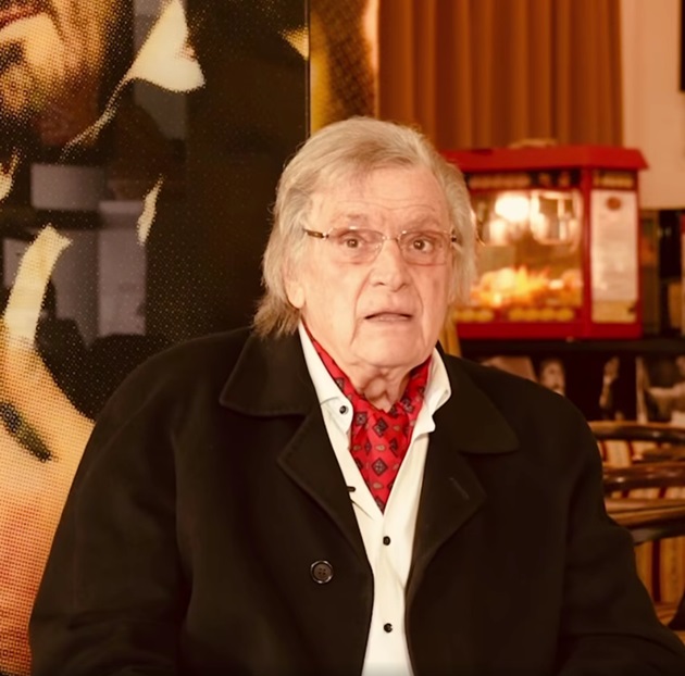 Florin Piersic la 89 de ani, în pulover alb, cu palton negru pe deasupra și eșarfă roșie la gât