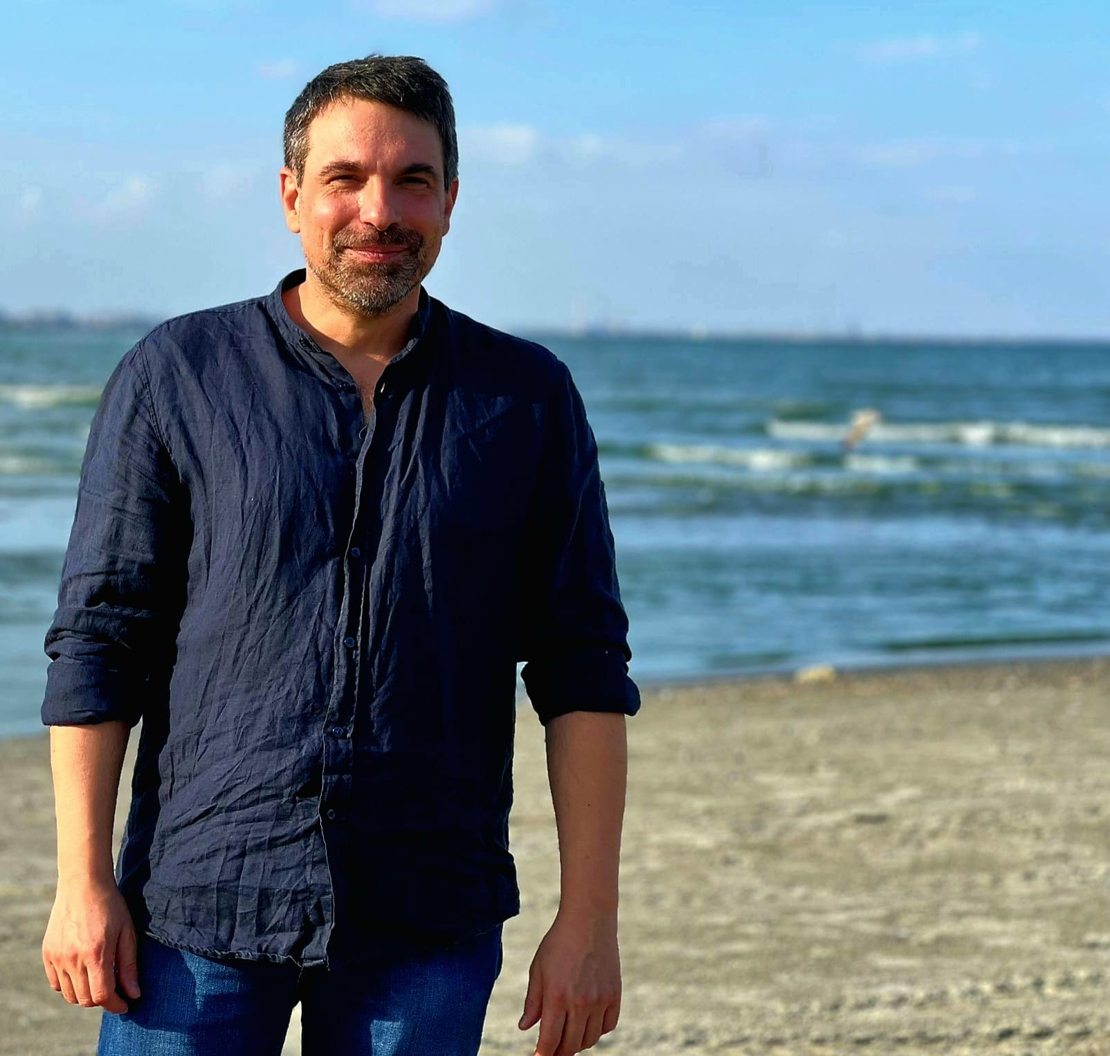 Alexandru Papadopol zâmbitor pe plajă, în blugi și cămașă bleumarin