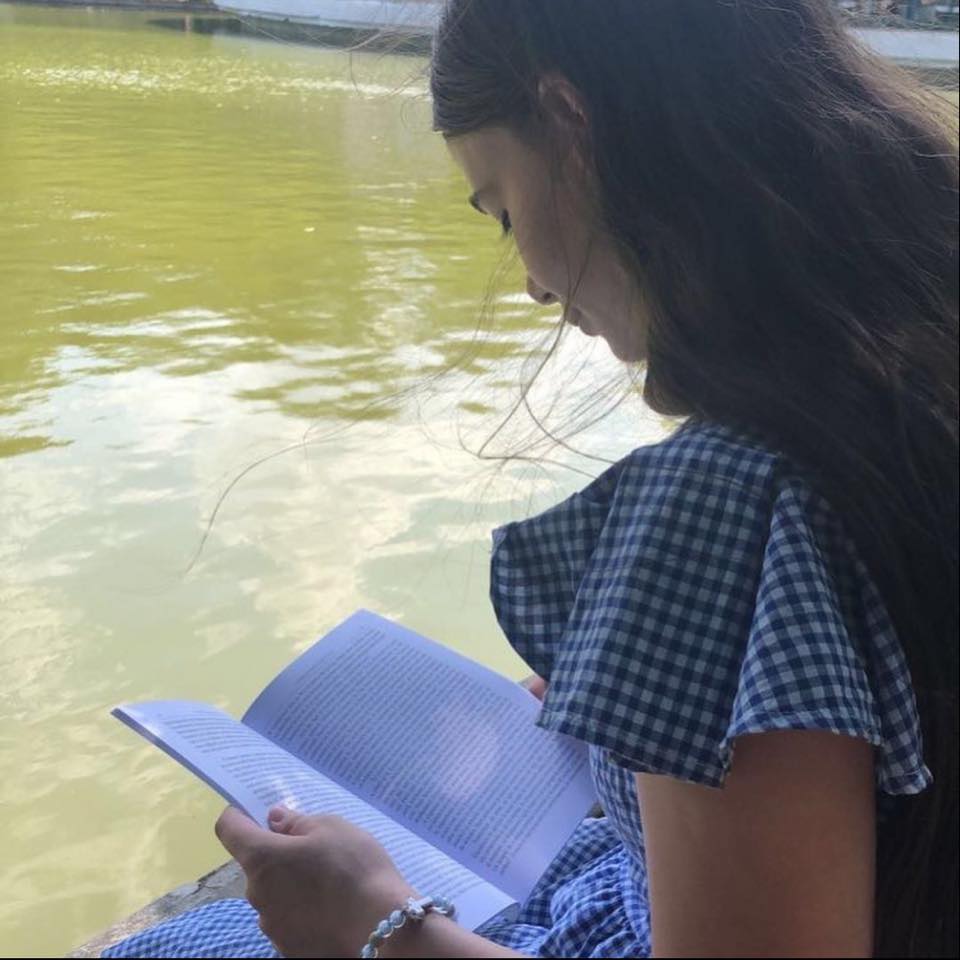 Eliza Iliescu stând pe malul unui lac și citind o carte