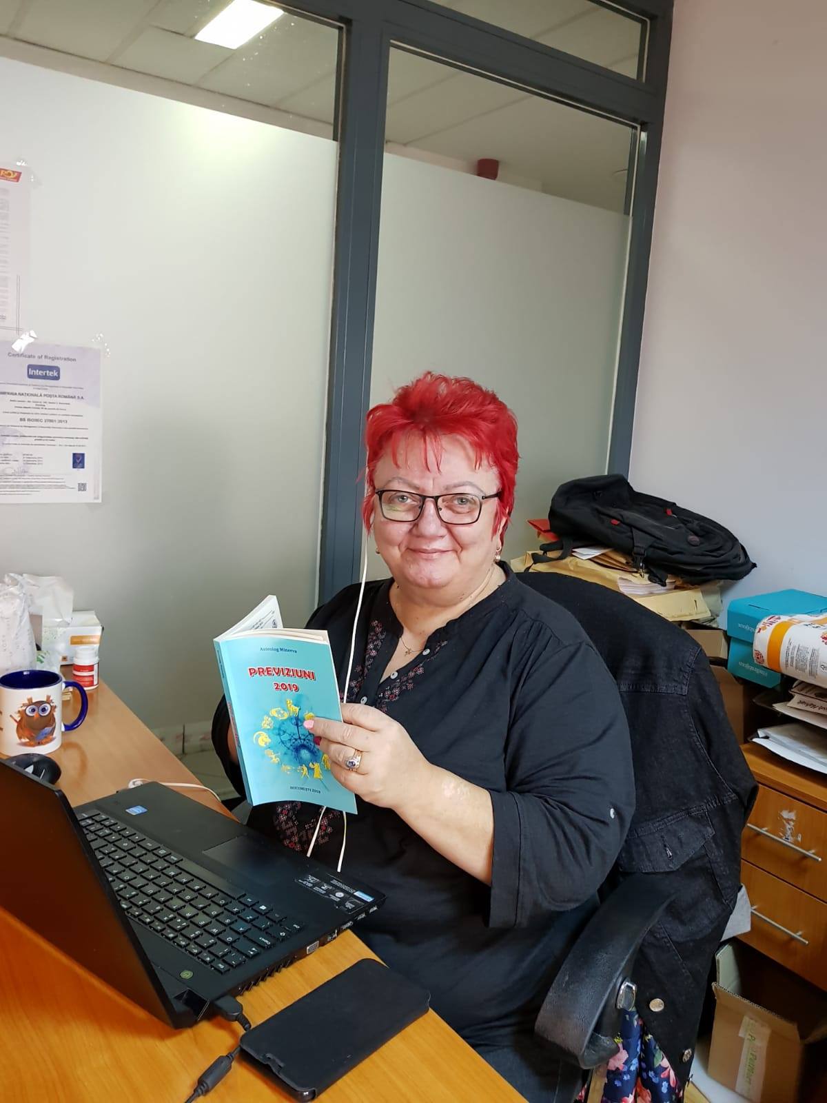 Minerva stând la birou și ținând în mână cartea cu previziunile ei pentru anul 2019