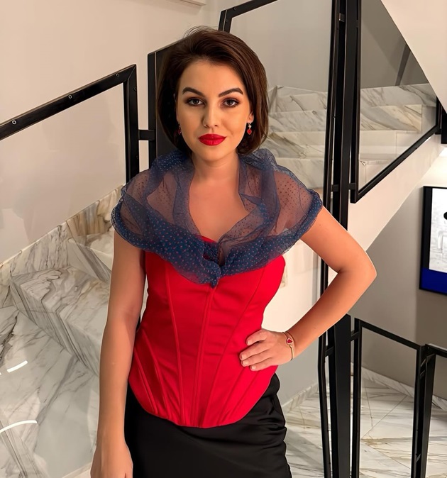 Romina Gingasu îmbrăcată în roșu și negru, cu i eșarfă albastră pe umeri, stând cu o mână în șold