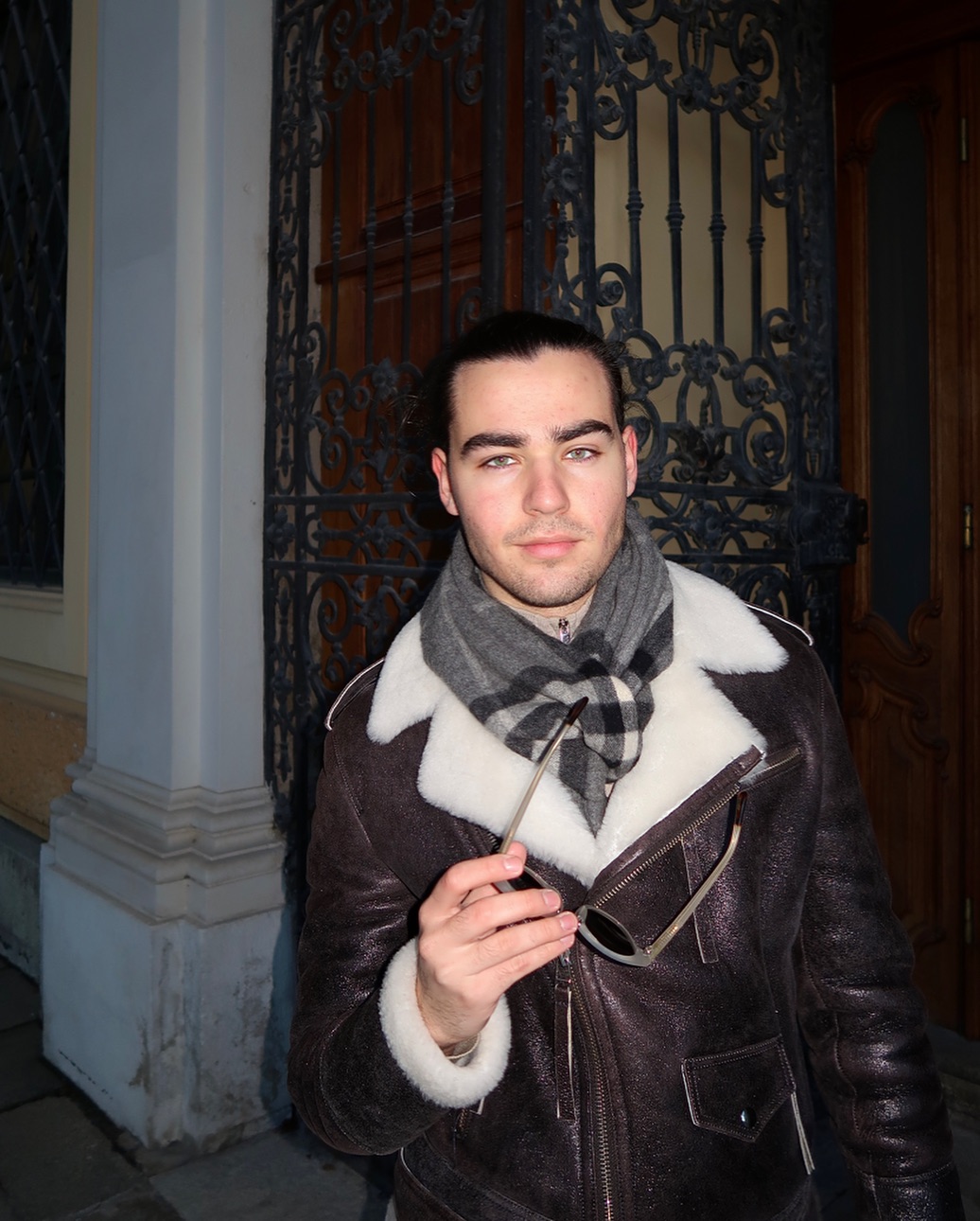 Radu Ștefan Bănică îmbrăcat într-o geacă de piele cu guler de blană, purtând la gât o eșarfă în carouri și ținând în mână o pereche de ochelari de soare