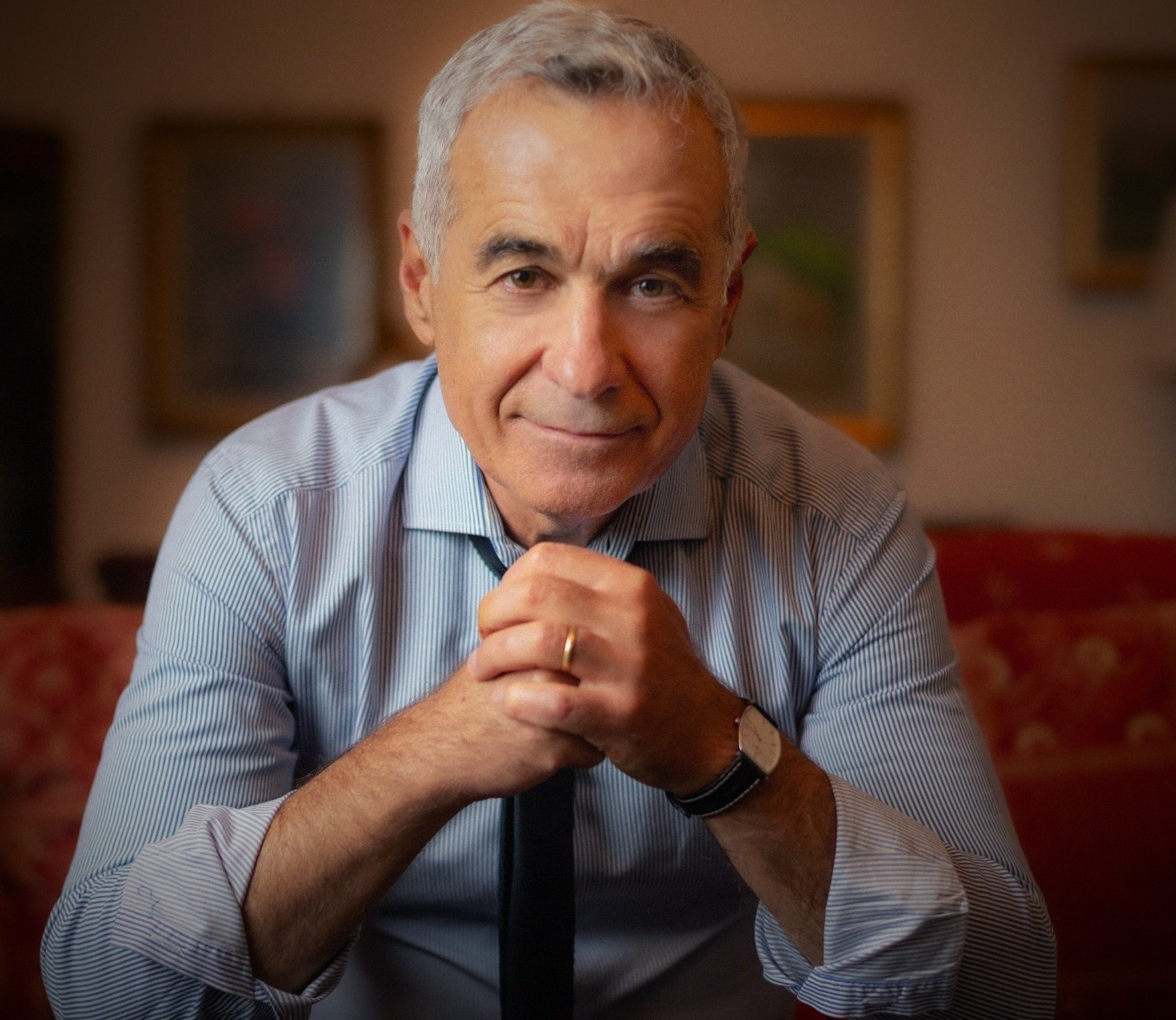 Călin Georgescu într-o cămașă cu dungi, stând pe canapea cu mâinile împreunate