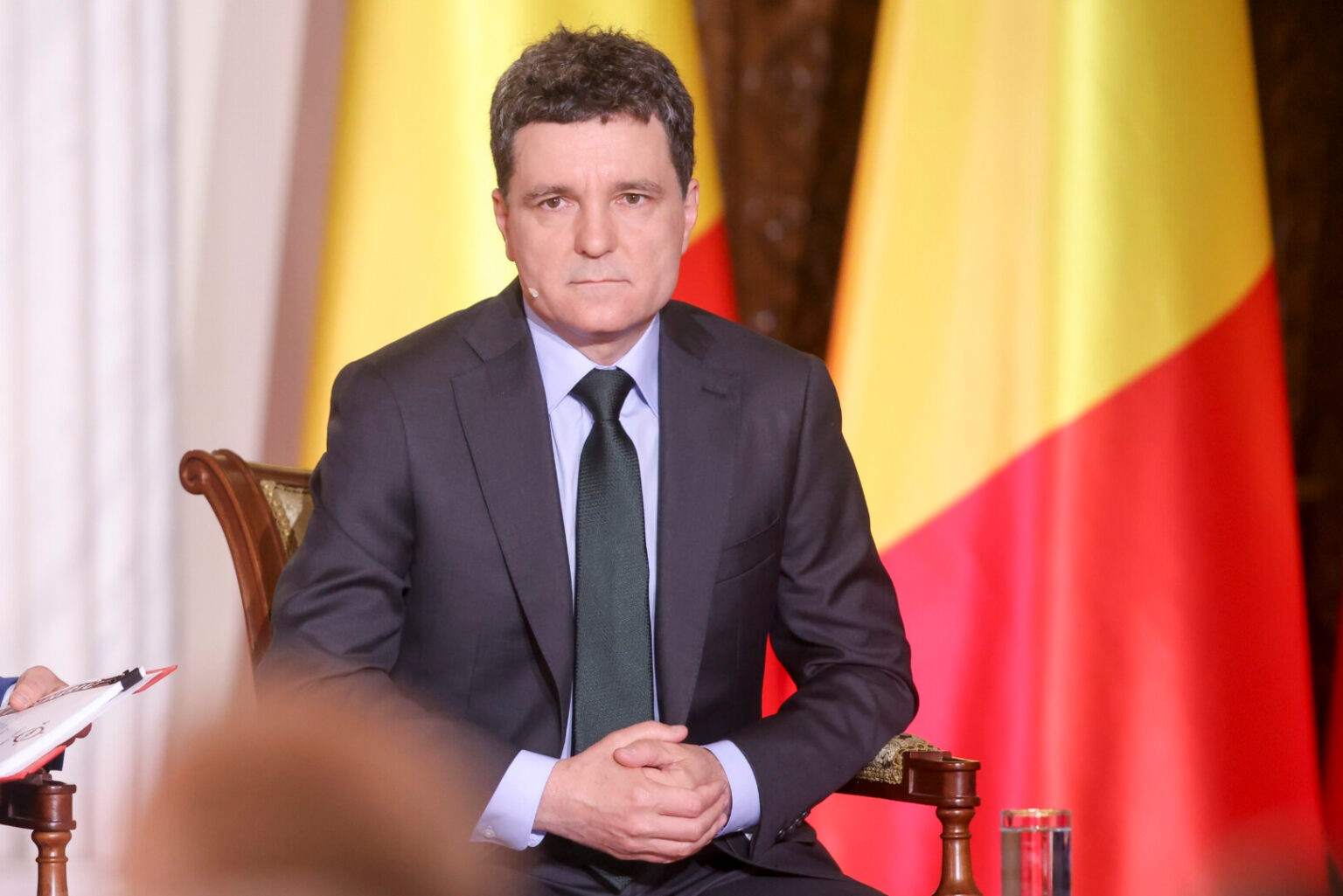 Nicușor Dan stând serios în fața steagului României, îmbrăcat în costum negru și cămașă deschisă la culoare