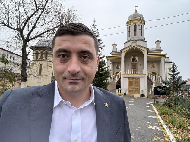 George Simion într-o cămașă albă, cu sacou gri și insigna României în piept, stând în fața unei biserici