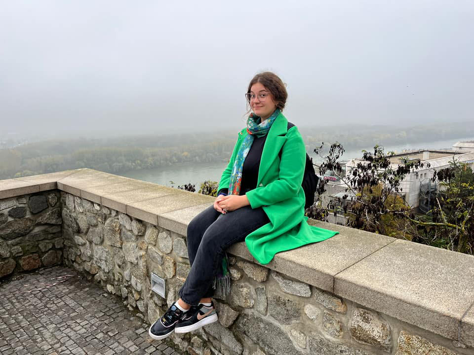 Shara Ionășchiță într-o excursie, îmbrăcată în jeanși negri și cardigan verde, purtându-și rucsacul în spate