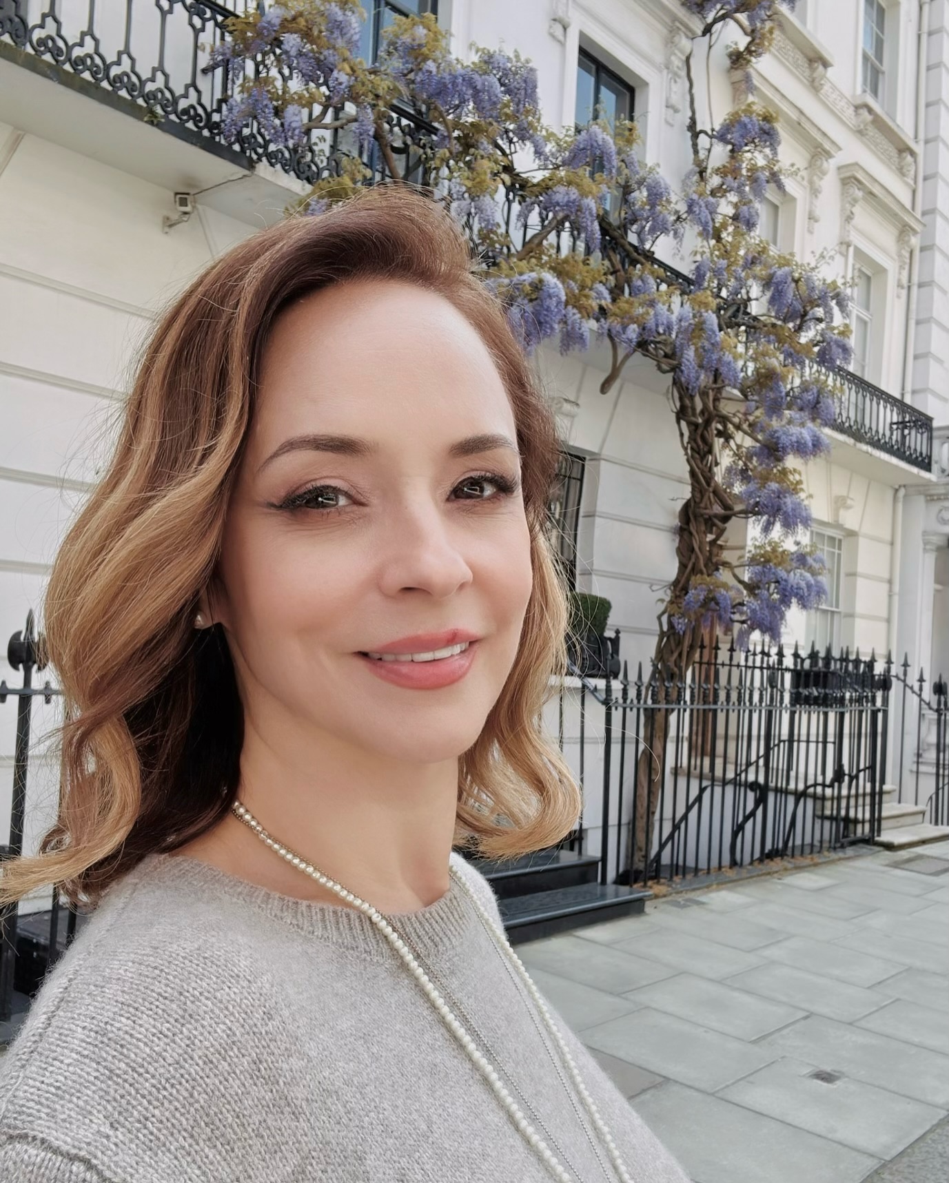 Andreea Marin zâmbitoare, în fața unei clădiri care are pe balcon o floare curgătoare