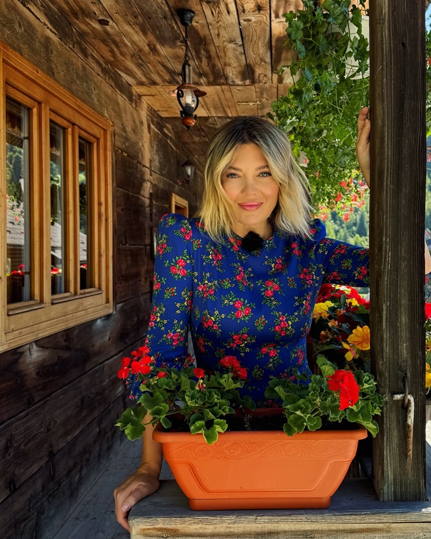 Gina Pistol într-o rochie albastră cu flori roșii, stând pe veranda unei case din lemn