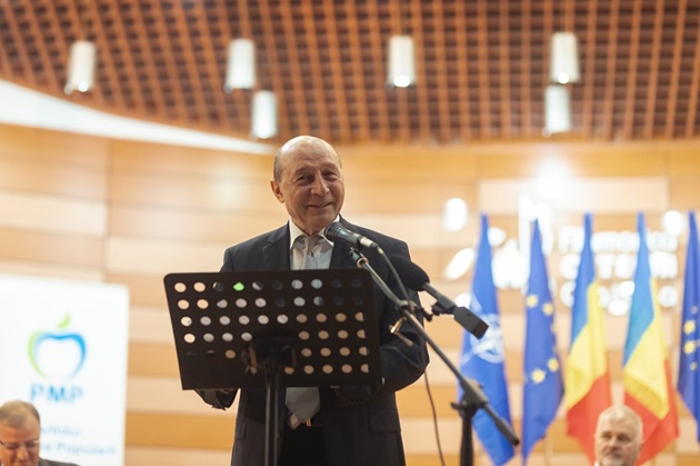 Traian Băsescu zâmbitor, îmbrăcat în costum negru, vorbind la microfon în fața colegilor săi politicieni