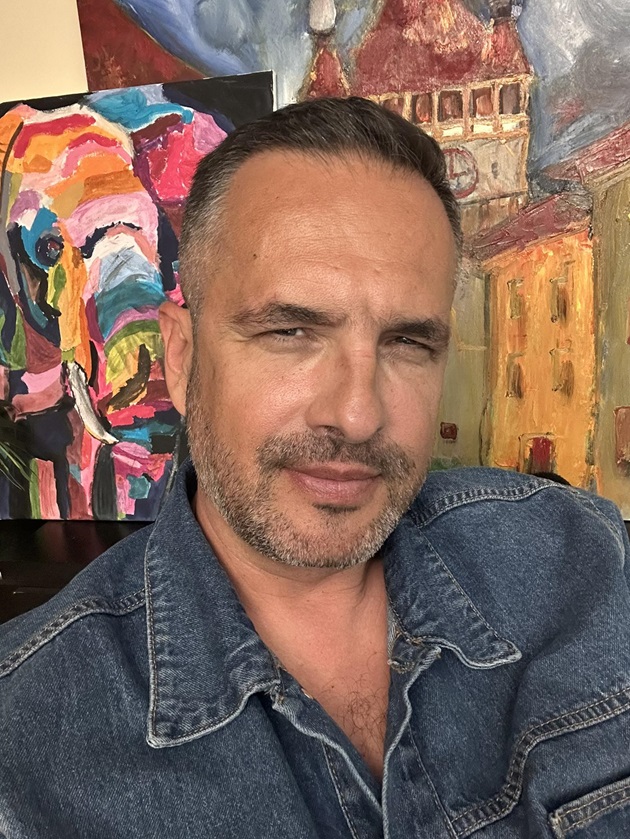 Mădălin Ionescu îmbrăcat într-o geacă de blugi, făcându-și un selfie în fața unor tablouri abstracte