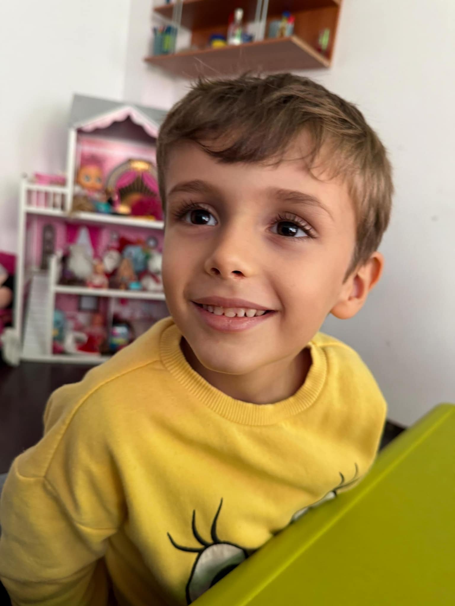 Geo, fiul cel mic al Iuliei Sabbagh și al lui Chistian Sabbagh, zâmbitor, stând la o masă verde, având în spate un raft plin cu jucării