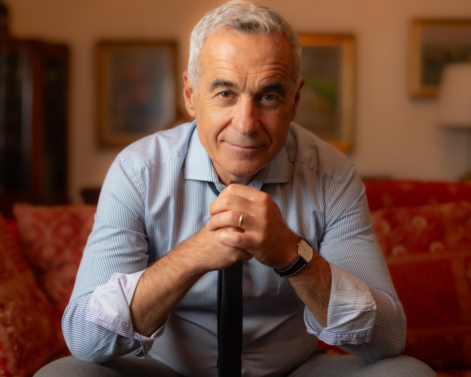 Călin Georgescu zâmbitor, stând cu mâinile împreunate sub bărbie, în timp ce era îmbrăcat în cămașă cu dungi și purta o cravată închisă la culoare
