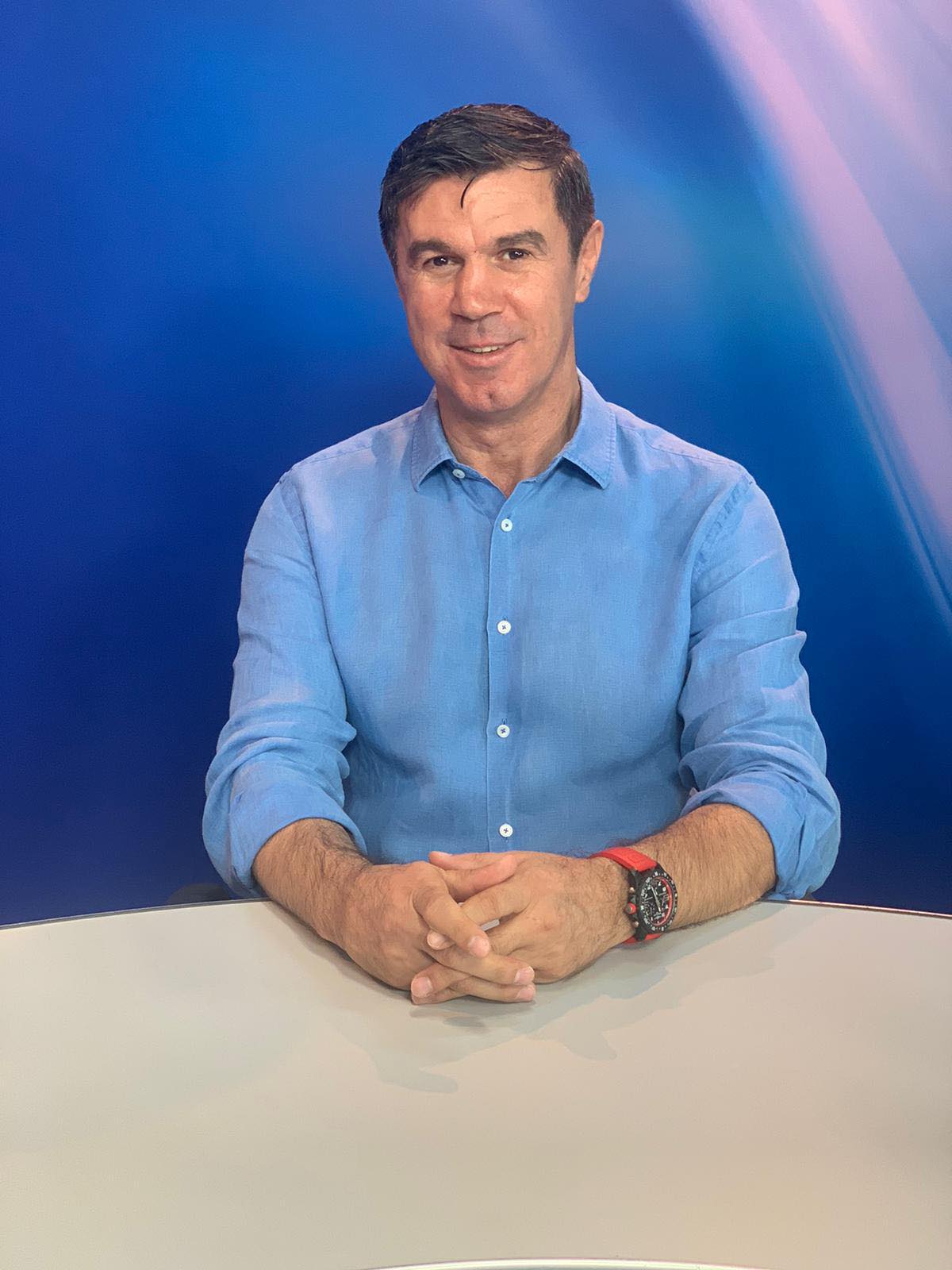 Mihai Leu zâmbitor, îmbrăcat într-o cămașă bleu, stând cu mâinile încrucișate pe o masă albă