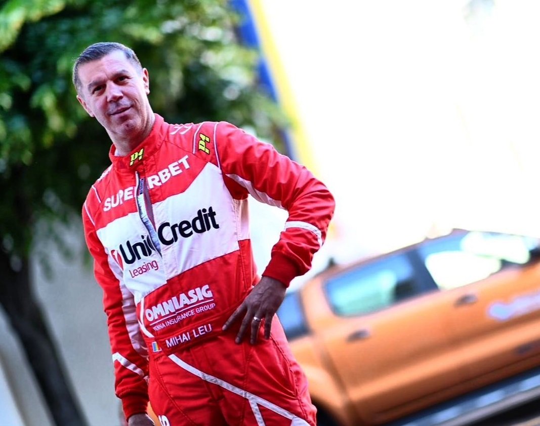 Mihai Leu în echipamentul în care participa la raliuri, o salopetă roșie cu numele sponsorilor pe ea, stâns cu mâna în șold, lăsând verigheta la vedere