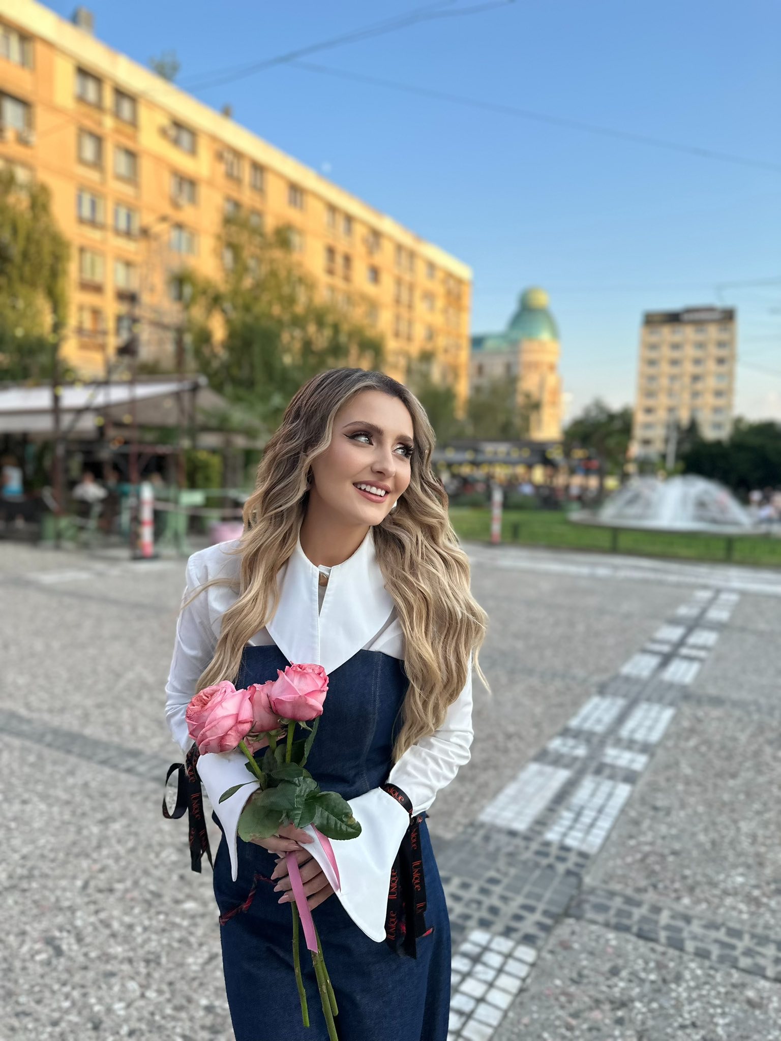 Alina Sorescu îmbrăcată în cămașă albă cu mâneci clopot și sarafan de blugi, ținând în mâini un buchet de trandafiri roz