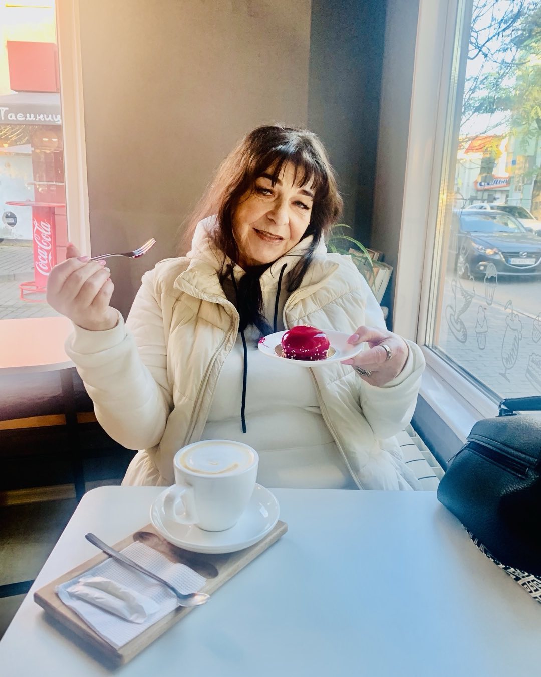 Alina Starukh îmbrăcată în alb și crem, stând la masa unei cafenele, savurând o cafea și o prăjitură