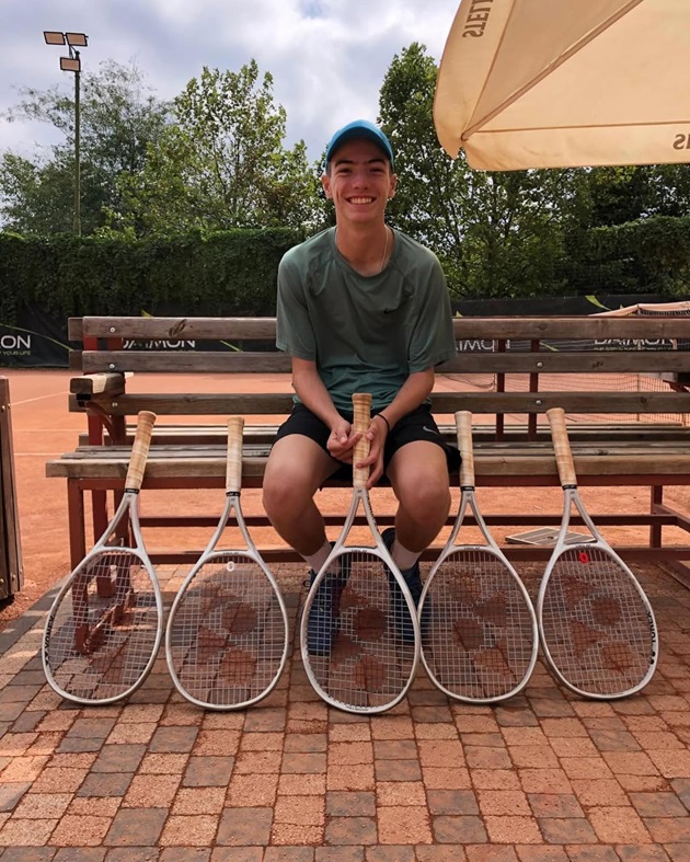Andrei Capatos, fiul lui Dan Capatos, stând pe o bancă pe marginea terenului de tenis, fiind înconjurat de palete de tenis