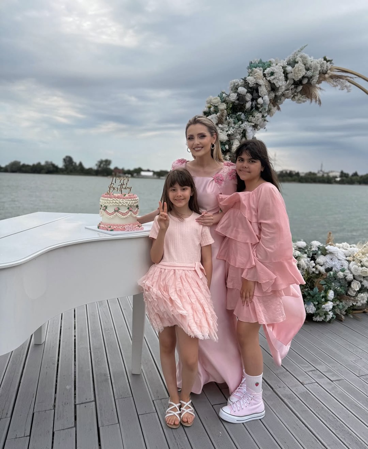 Alina Sorescu pe un ponton, la filmarea unui videoclip, îmbrăcată într-o rochie lungă, roz, alături de fiicele sale, Raisa și Carolina, care s-au îmbrăcat asortat cu ea