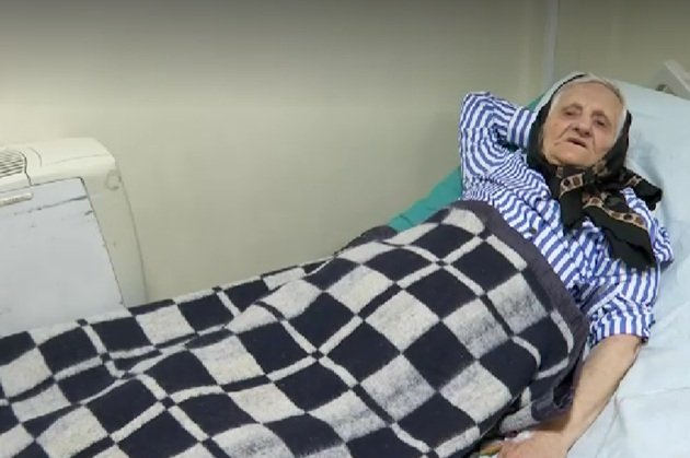 Aurica, femeia de 84 de ani din Iași abandonată de familie în spital, stând întinsă în pat, cu o mână sub cap și învelită cu o pătură în carouri