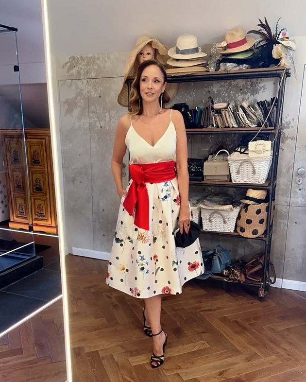Andreea Marin cu o mână în buzunar, îmbrăcată într-o rochie albă cu flori, la care și-a pus sandale negre și o poșetă neagră